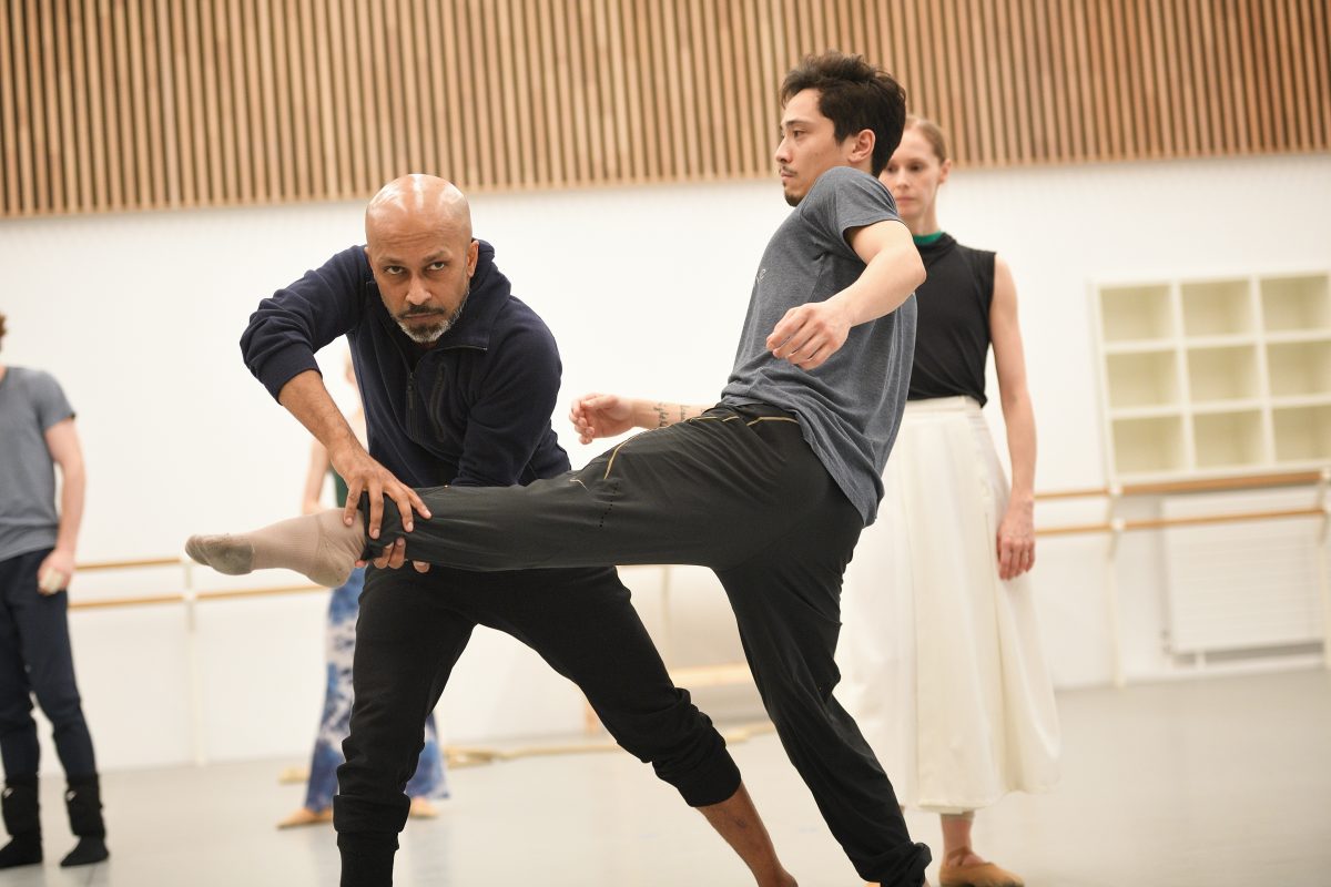 Choreographer assisting a male dancer in a rehearsal studio, with another dancer observing in the background. The studio has modern lighting, wooden panel accents, and ballet barres along the walls.