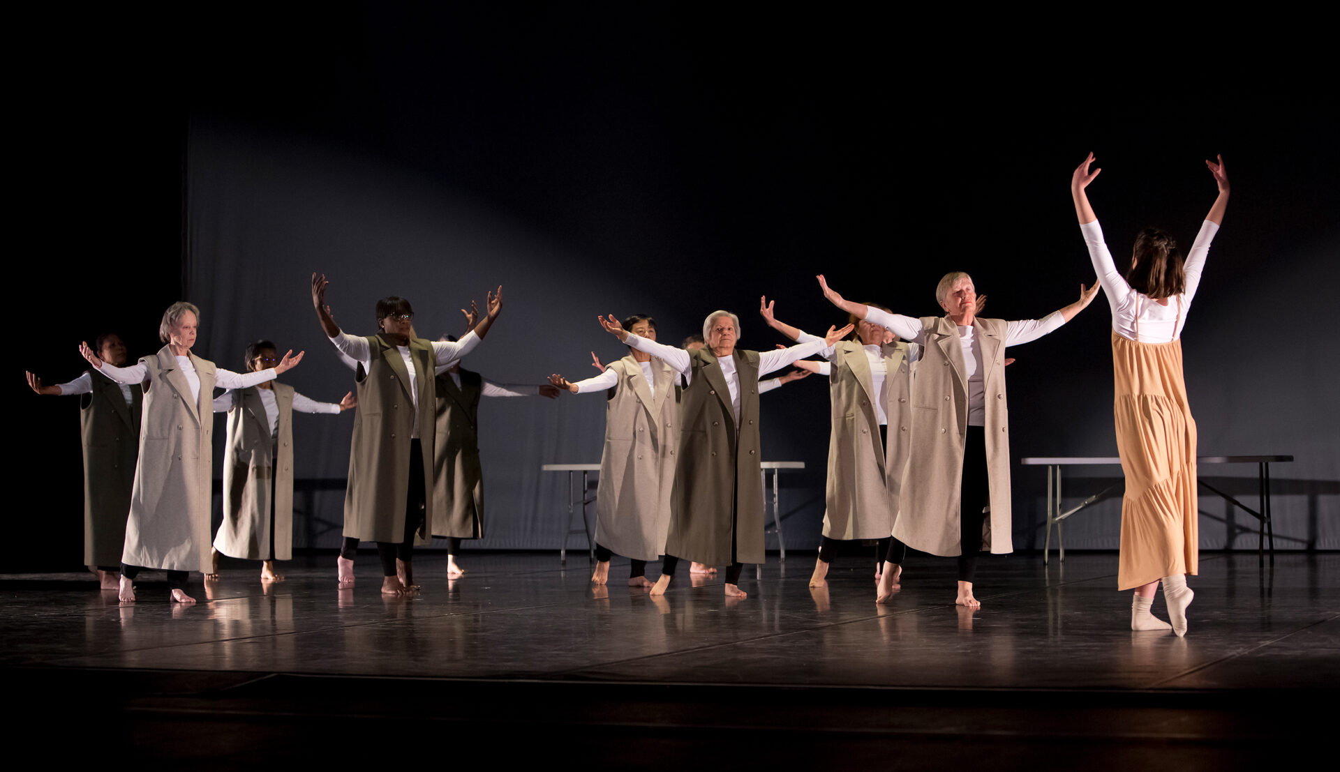 A dance group of older adults is performing on stage, all dressed in long, sleeveless beige coats over white tops and black pants. The dancers are barefoot and moving in unison with their arms outstretched, following the lead of a younger dancer in a flowing peach-colored dress who stands at the front. The background is dark.