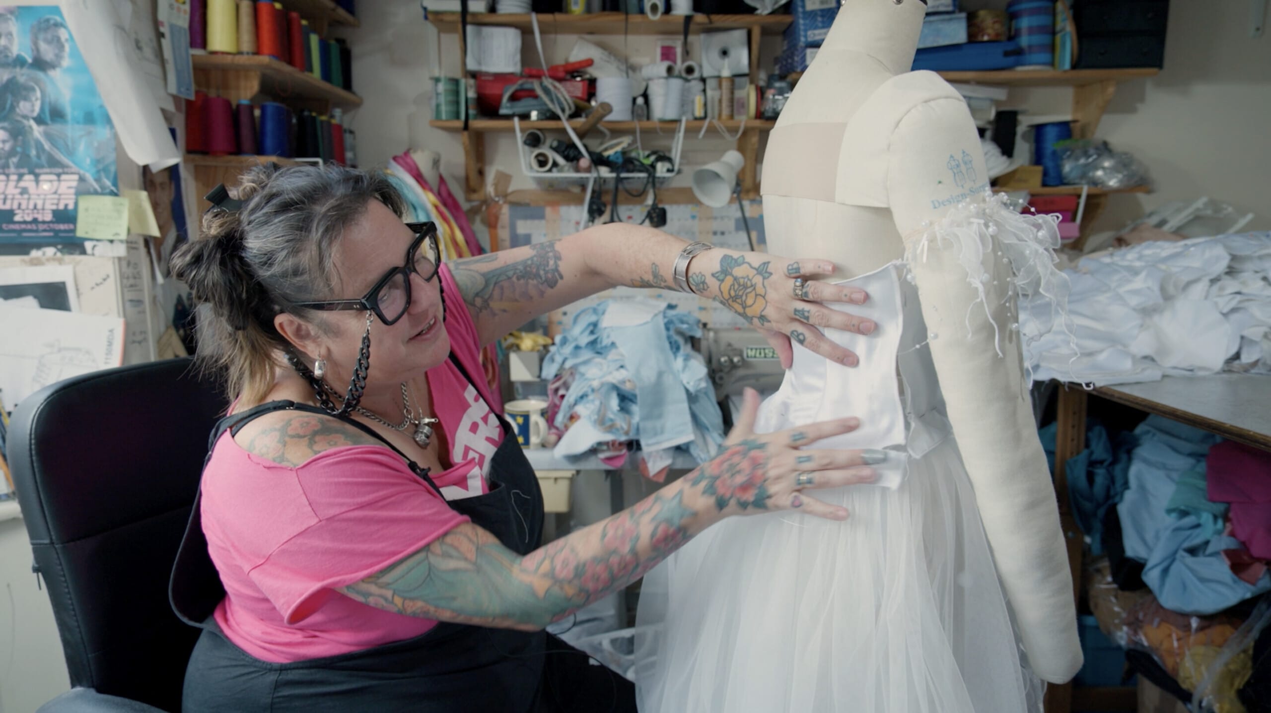 A woman with tattoos and glasses is working on a white dress on a mannequin in a cluttered sewing studio. She is adjusting the bodice of the dress, which is made of a shiny white fabric, while the skirt is made of tulle. The workspace is filled with sewing supplies, fabric, and tools, with colourful thread spools.