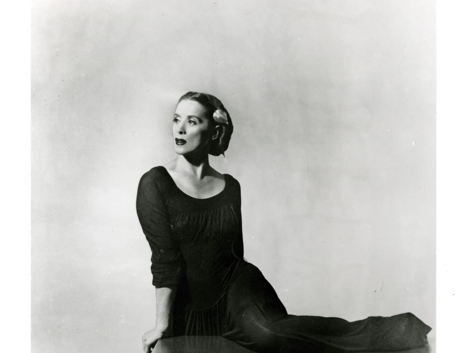 A woman in a long, dark dress sits gracefully on a wooden table, one leg extended along its surface. She looks to the side with a composed expression, her hair styled neatly with a decorative accessory.